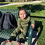 child, smiling, bench, outdoor, park, grass, backpack, green_clothing, dinosaur_pattern, sneakers, sunlight, happy, casual_wear, concrete, trees, daytime, person, seat, relaxing, nature
