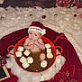 baby, santa_hat, marshmallows, red_pot, christmas, holiday_decor, cotton_fluff, wooden_floor, gift_boxes, festive, infant, cute, holiday_theme, holiday_lighting, red_blanket, merry_christmas, face, person, indoors, celebration