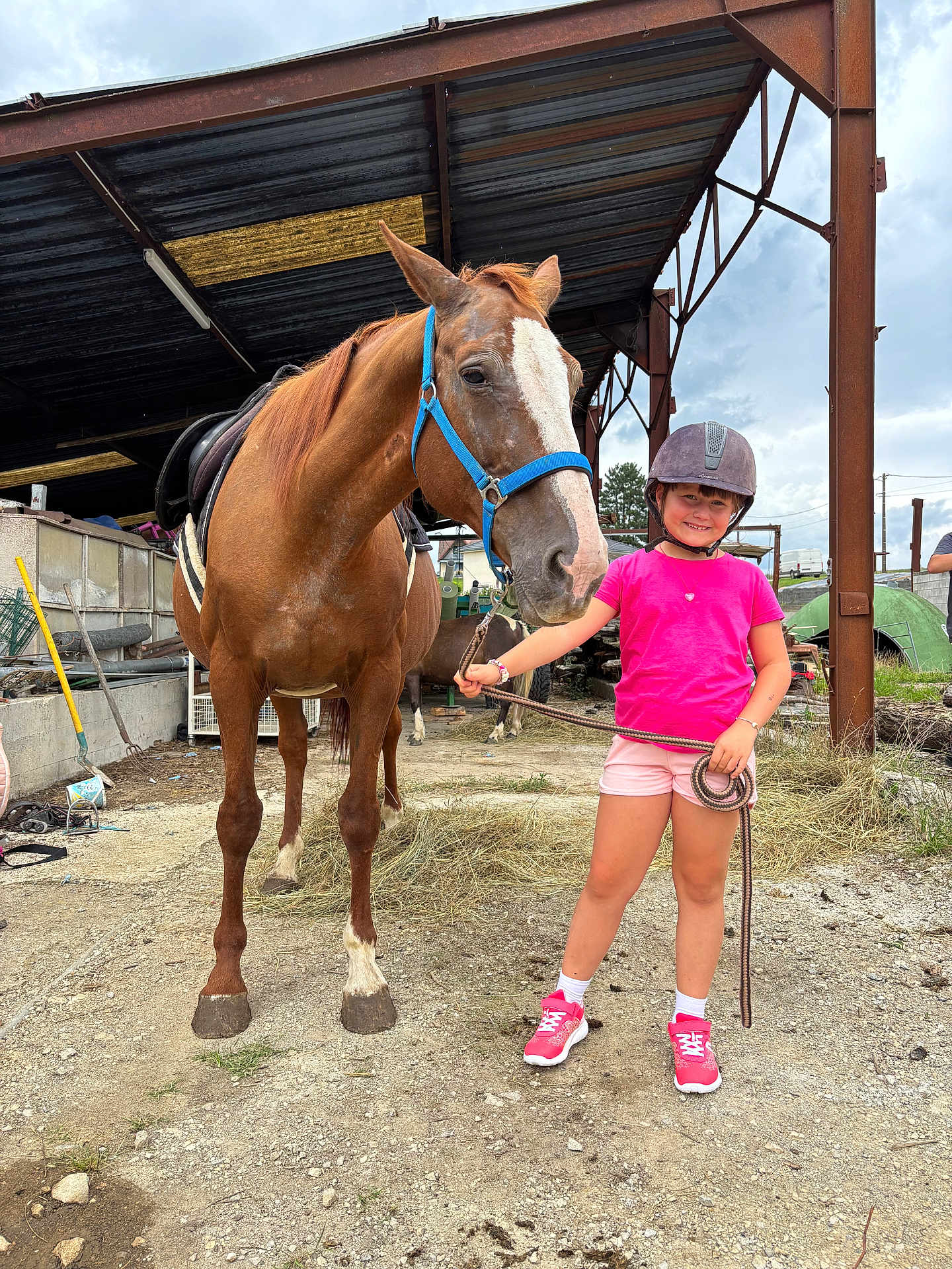 élysée a rejoint le concours — aidez-le/la à gagner de superbes lots ! child, horse, animal, helmet, pink_shirt, shorts, reins, farm, outdoor, smiling, dirt_ground, metal_shelter, grass, sky, casual_clothing, footwear, rope, sunlight, young_person, pet