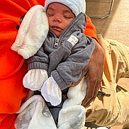 Zakkai is registered to the contest to win money with this photo: baby, sleeping, infant, blanket, clothing, hood, hand, person, orange_sweatshirt, gray_jacket, white_hat, white_mittens, warm, cozy, indoor, wood_floor, chair, toddler, peaceful, sunlight