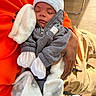 baby, sleeping, infant, blanket, clothing, hood, hand, person, orange_sweatshirt, gray_jacket, white_hat, white_mittens, warm, cozy, indoor, wood_floor, chair, toddler, peaceful, sunlight