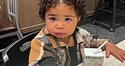 Kaliyah is registered to the contest to win money with this photo: child, toddler, curly_hair, juice_box, honest_kids, plaid_jacket, table, chair, indoor, wooden_wall, curious_expression, black_chair, high_chair, person, young_child, drink, juice, furniture, casual, dining