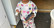 Kaliyah is registered to the contest to win money with this photo: carpet, child, colorful, curly_hair, cute, doorway, expression, footwear, home, indoor, laundry_basket, mat, pacifier, pajamas, person, playful, pout, smartphone, standing, toddler