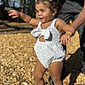 child, toddler, white_romper, bows, hair_accessories, walking, playground, wood_chips, sunlight, smile, supportive_adult, sandals, outdoor, happy, person, cute, curly_hair, skin, summer, fun