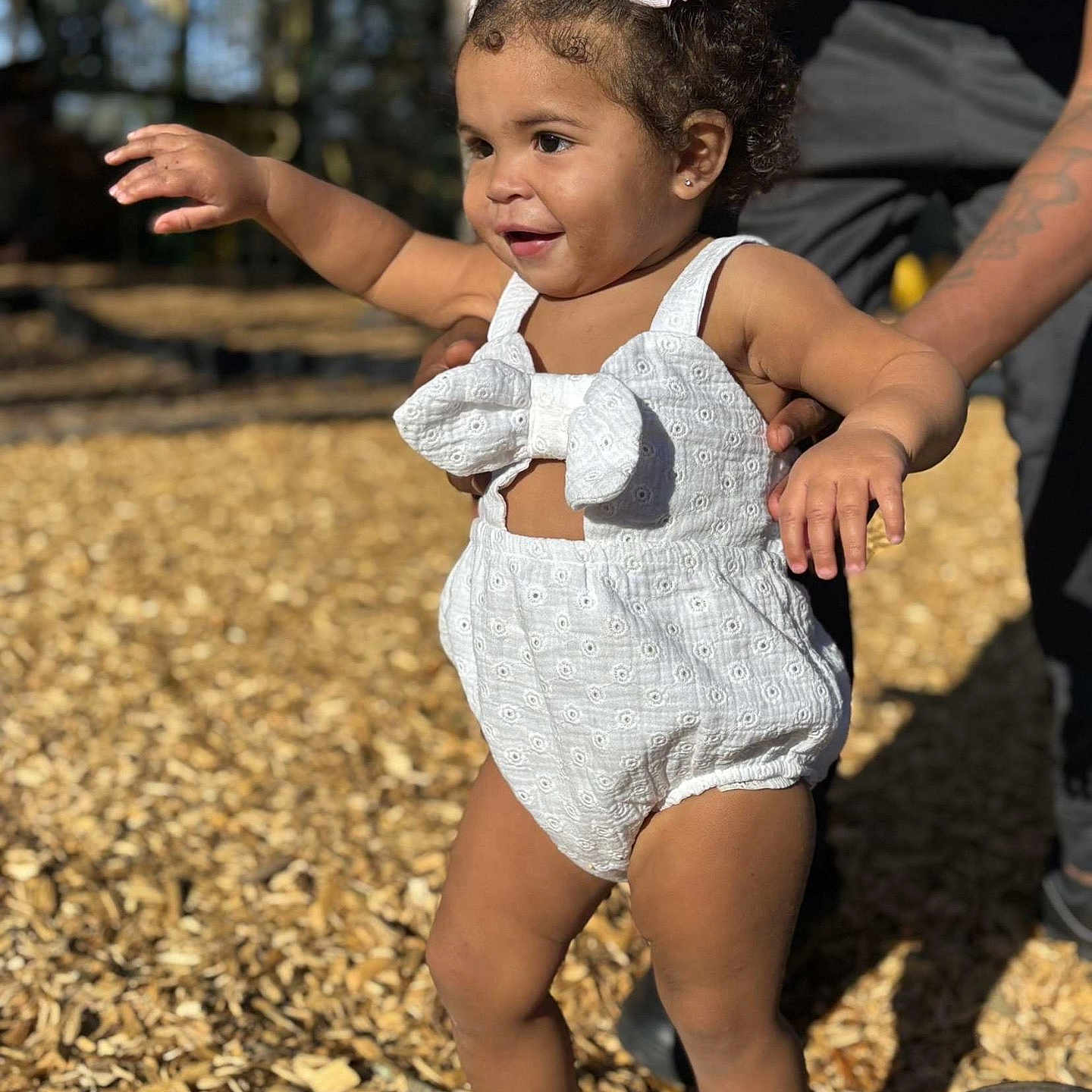 Kaliyah joined the competition — help win amazing prizes! child, toddler, white_romper, bows, hair_accessories, walking, playground, wood_chips, sunlight, smile, supportive_adult, sandals, outdoor, happy, person, cute, curly_hair, skin, summer, fun