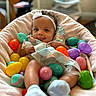 baby, child, headband, easter_eggs, colorful, plastic_eggs, soft_chair, blanket, playful, tongue_out, indoors, cute, smiling, infant, cozy, person, clothing, socks, portrait, happy