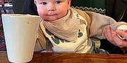 Teddy is registered to the contest to win money with this photo: baby, child, cup, table, wooden_table, person, hand, clothing, bib, indoor, portrait, face, smile, holding, seated, adult, colorful, cozy, family, restaurant