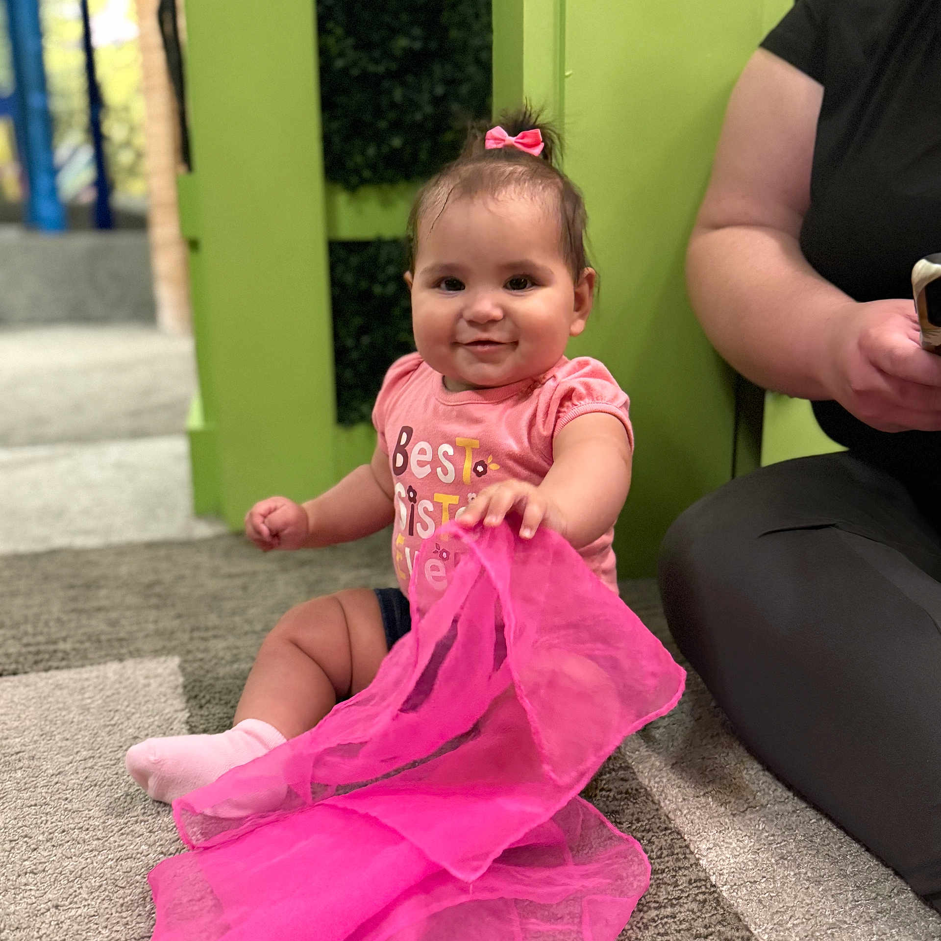 Aelfwynn is registered to the contest to win money with this photo: baby, child, pink_bow, pink_shirt, sitting, carpet, fabric, playful, smiling, indoor, person, adult, green_wall, toy, cute, happy, girl, floor, casual_clothing, holding