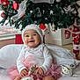 baby, infant, smiling, christmas_tree, ornament, bauble, pink_tutu, santa_hat, white_top, pompom_sock, present, holiday_decor, indoors, pillow, bed, festive, cute, portrait, happy, toy