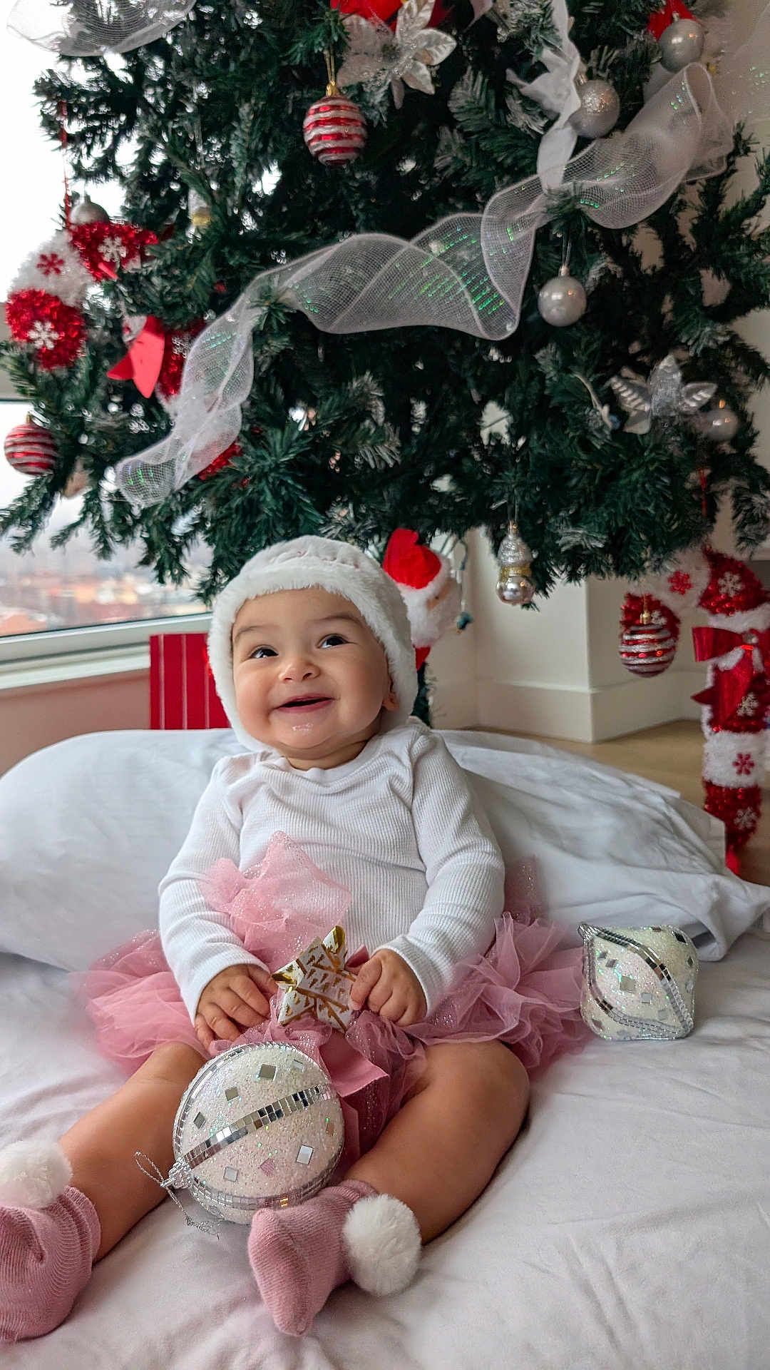 Mirana joined the competition — help win amazing prizes! baby, infant, smiling, christmas_tree, ornament, bauble, pink_tutu, santa_hat, white_top, pompom_sock, present, holiday_decor, indoors, pillow, bed, festive, cute, portrait, happy, toy