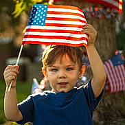 Luca is registered to the contest to win money with this photo: child, boy, american_flag, flag, outdoor, daylight, grass, tree, patriotic, celebration, holiday, summer, casual_clothing, blue_shirt, young, person, nature, holding, festive, portrait