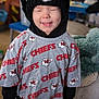 child, smiling, closed_eyes, hat, knit_hat, shirt, chiefs, indoor, cozy, happy, young_child, casual_clothing, portrait, person, cute, black_clothing, gray_shirt, soft_texture, background, standing