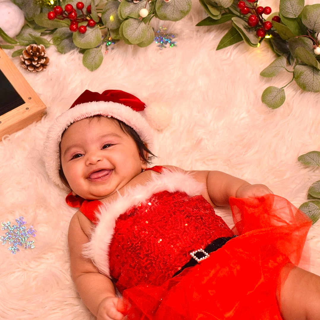 Raavya is registered to the contest to win money with this photo: baby, celebration, child, christmas, christmas_hat, cute, festive, fluffy_rug, greenery, happy, holiday, indoors, infant, pine_cone, portrait, red_dress, smile, smiling, snowflake_decoration, toddler