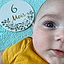 baby, child, face, eye, skin, clothing, yellow_shirt, milestone_card, text, blue_blanket, closeup, portrait, infant, cute, young_child, human, laying_down, expression, indoor, soft_texture
