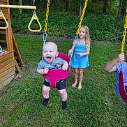 Walker is registered to the contest to win money with this photo: baby, child, swing, playground, grass, trees, outdoor, play, happy, smiling, toddler, girl, barefoot, blue_dress, wooden_play_structure, fun, summer, nature, yellow_chains, greenery