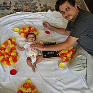 Mira is registered to the contest to win money with this photo: baby, man, infant, parent, garland, marigold, flower_garland, paint, paint_bowl, white_sheet, rug, floor, name_board, portrait, smile, hand, eyes, home, casual_clothing, indoor