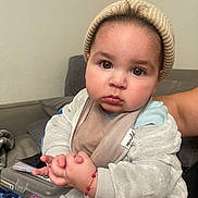 Ezra is registered to the contest to win money with this photo: baby, child, indoor, hat, beanie, clothing, hands, face, expression, person, cute, portrait, warm, soft, jacket, couch, bracelet, skin, eyes, sitting
