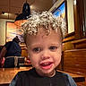 child, toddler, curly_hair, smile, restaurant, wooden_table, indoor, person, face, lighting, waiter, background, artwork, wood_paneling, casual, dining, curly_hair_boy, young_child, happy, portrait