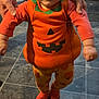 Bonnie is registered to the contest to win money with this photo: baby, child, pumpkin_costume, orange_clothing, smiling, standing, tattooed_arms, floor, tiles, indoor, person, cute, holidays, halloween, happy, support, hair_tie, legs, socks, facial_expression