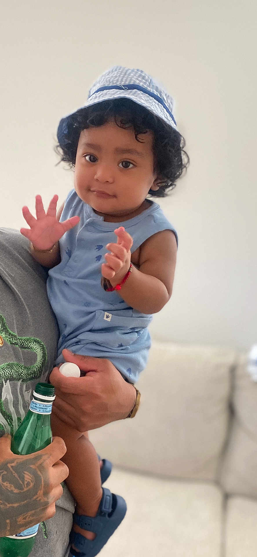 Denilson is registered to the contest to win money with this photo: toddler, child, hat, blue_clothing, curly_hair, person, hand, tattoo, bottle, adult, indoor, couch, footwear, sandals, bracelet, smiling, holding, portrait, casual, cute