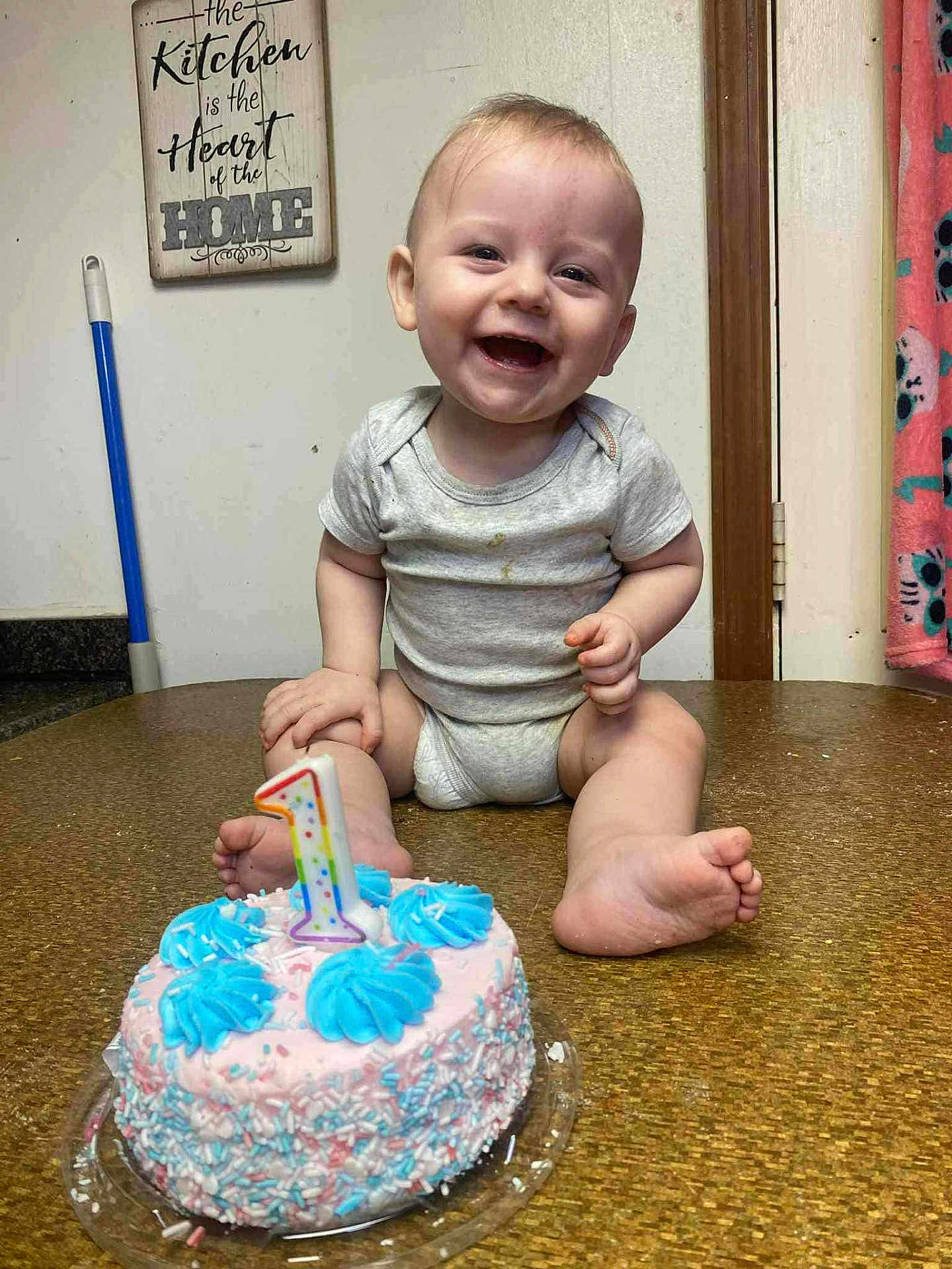 Levi is registered to the contest to win money with this photo: baby, infant, smiling, cake, number_candle, first_birthday, onesie, sitting, toes, frosting, sprinkles, table_top, kitchen_sign, indoor, doorway, towel, broom, celebration, happy, portrait