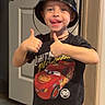 Hudson is registered to the contest to win money with this photo: blue_eyes, bucket_hat, cars, child, costume, doorway, funny_face, happy, hat, home, indoors, lightning_mcqueen, photograph, portrait, smiling, standing, t_shirt, thumbs_up, toddler, toy