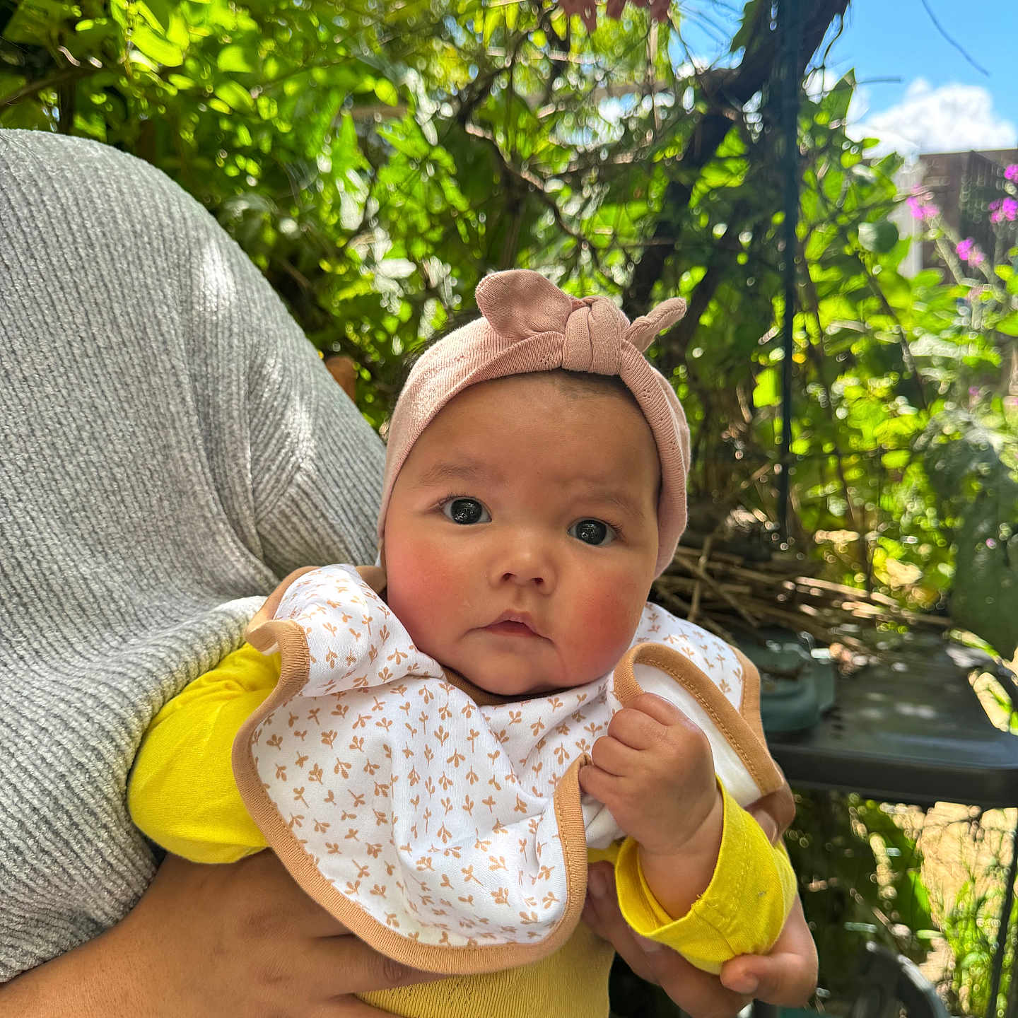 Meï-Li a rejoint le concours — aidez-le/la à gagner de superbes lots ! adult_hand, baby, bib, child, chubby_cheeks, clothing, curious, cute, denim_pants, face, garden, greenery, headband, holding, infant, outdoor, person, portrait, sunlight, yellow_shirt