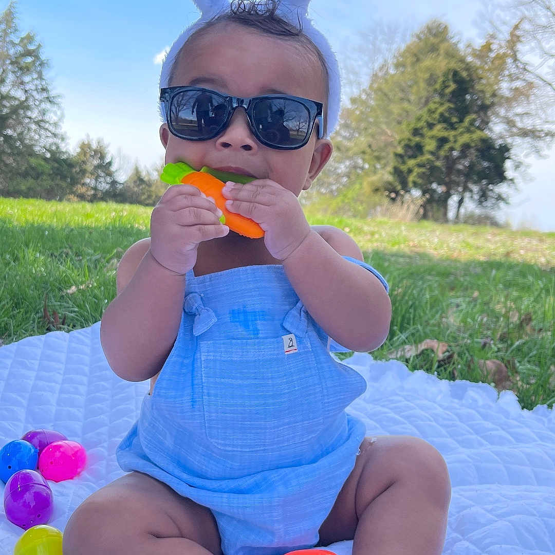 Amani is registered to the contest to win money with this photo: accessories, baby, bodypart, face, finger, food, glasses, grass, hand, head, nature, outdoors, park, person, photography, plant, portrait, summer, sunglasses, tree