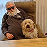 Pépette participe au concours pour gagner de l'argent avec cette photo : elderly_man, white_beard, sunglasses, dog, fluffy_dog, chair, curtains, table, indoor, smiling_dog, casual_clothing, seated, pet, companion, relaxed, home, portrait, cozy, hand_on_dog, beard