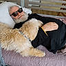 elderly_man, dog, bed, cuddling, indoor, fan, sunglasses, white_beard, light_fur_dog, blanket, wooden_floor, chair, photographs, relaxation, casual_clothing, home, resting, affection, pet, companion