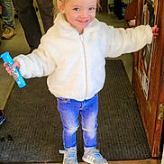 Hazel is registered to the contest to win money with this photo: toddler, child, smiling, white_jacket, blue_jeans, sneakers, indoors, doorway, carpet, holding_snack, hair_bow, happy, person, standing, floor, wooden_door, shelves, casual_clothing, cute, portrait