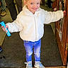 toddler, child, smiling, white_jacket, blue_jeans, sneakers, indoors, doorway, carpet, holding_snack, hair_bow, happy, person, standing, floor, wooden_door, shelves, casual_clothing, cute, portrait