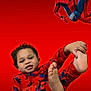 child, toddler, pajamas, spiderman, superhero, red_background, smile, foot, feet, playful, costume, masked_character, comic_character, portrait, pose, indoor, patterned_clothing, camera_looking, young_child, bright_color