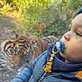 child, toddler, pacifier, pacifier_clip, jacket, zipper, tiger, animal, zoo, glass_barrier, wildlife, outdoor, foliage, portrait, close_up, curious, beanie, toy_lion, baby_accessory, bright_colors
