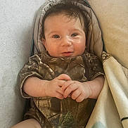 Grayson joined the competition — help win amazing prizes! baby, infant, smiling, camo_clothing, blanket, lying_down, cute, hand_gesture, indoors, portrait, child, young, cozy, soft_lighting, face, small_hands, happy, person, resting, warm
