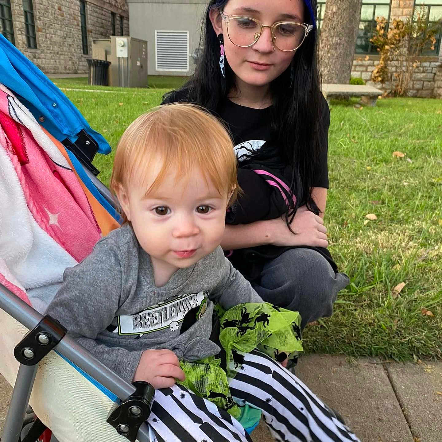 Selena is registered to the contest to win money with this photo: black_hair, blue_hair, building, casual_clothing, child, curly_hair, cute, daylight, girl, glasses, grass, outdoor, park, playing, sidewalk, sitting, striped_pants, stroller, toddler, young_person