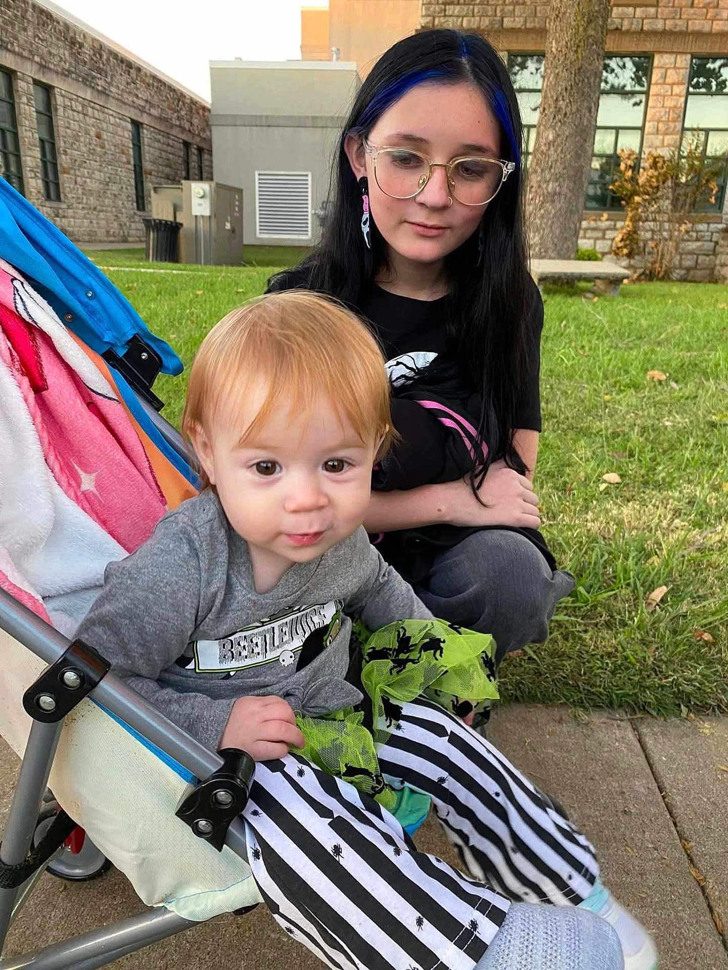 Selena is registered to the contest to win money with this photo: toddler, child, girl, stroller, glasses, outdoor, grass, building, sidewalk, black_hair, blue_hair, striped_pants, casual_clothing, young_person, playing, park, curly_hair, sitting, cute, daylight