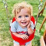 Samuel joined the competition — help win amazing prizes! toddler, child, baby, smiling, happy, curly_hair, blue_eyes, portrait, outdoor, playground, swing, red_swing, grass, summer, sitting, face, person, shorts, t_shirt, playful