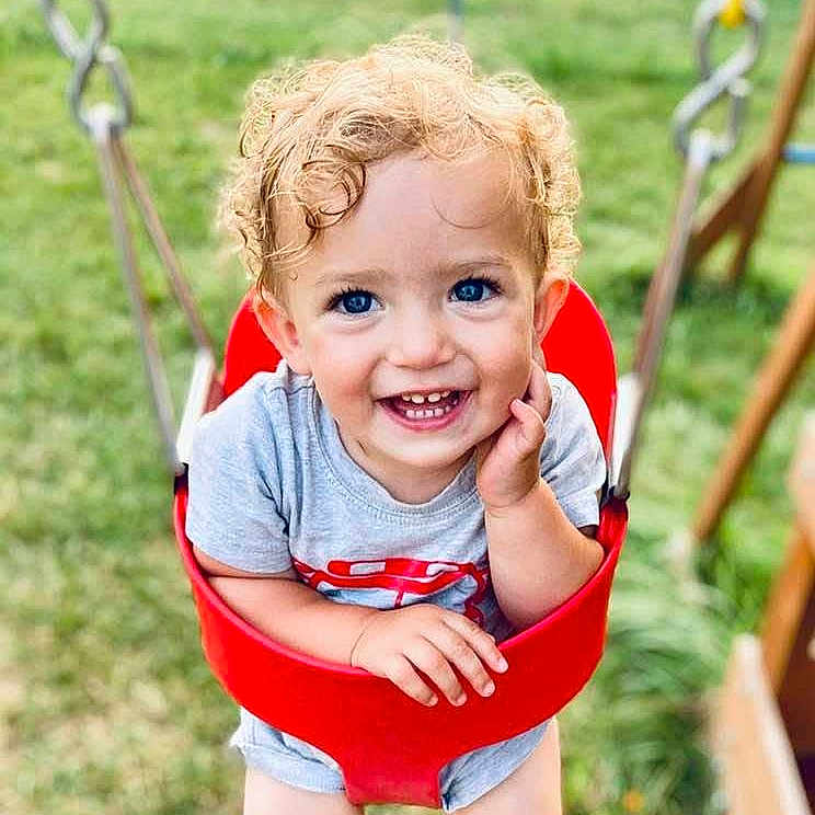 Samuel joined the competition — help win amazing prizes! toddler, child, baby, smiling, happy, curly_hair, blue_eyes, portrait, outdoor, playground, swing, red_swing, grass, summer, sitting, face, person, shorts, t_shirt, playful