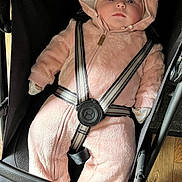 Olivia is registered to the contest to win money with this photo: baby, child, pink_onesie, stroller, baby_boy, infant, face, straps, footwear, floor, wood_floor, hood, cute, baby_clothing, safety_harness, indoor, seated, person, young_child, plush