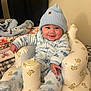 child, baby, infant, smiling, blue_beanie, hat, onesie, plush_chair, inflatable_seat, blanket, bedding, pillow, indoors, cozy, rosy_cheeks, adorable, sitting, hand, face, portrait