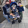 baby, boy, child, clothing, family, father, indoor, jacket, lobby, masked_person, portrait, shoes, sitting, smiling, standing, stroller, tile_floor, toddler, travel, wheelchair_accessible