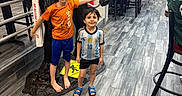 Sulaiman is registered to the contest to win money with this photo: children, boys, child, smiling, portrait, gorilla_statue, restaurant, indoor, soccer_jersey, orange_shirt, blue_shorts, football, standing, posing, playful, high_five, furniture, chairs, wood_floor, american_flag