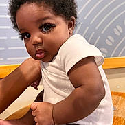 Chess is registered to the contest to win money with this photo: baby, infant, child, cute, portrait, sitting, wooden_surface, curly_hair, expression, pout, skin, white_clothing, indoor, person, young_child, closeup, human, face, arm, leg