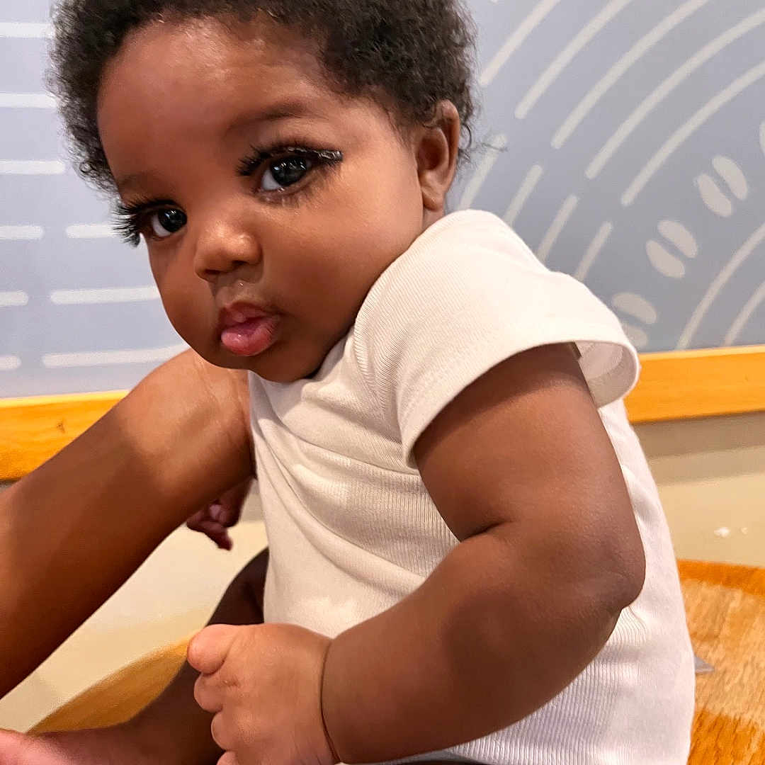 Chess is registered to the contest to win money with this photo: arm, baby, child, closeup, curly_hair, cute, expression, face, human, indoor, infant, leg, person, portrait, pout, sitting, skin, white_clothing, wooden_surface, young_child