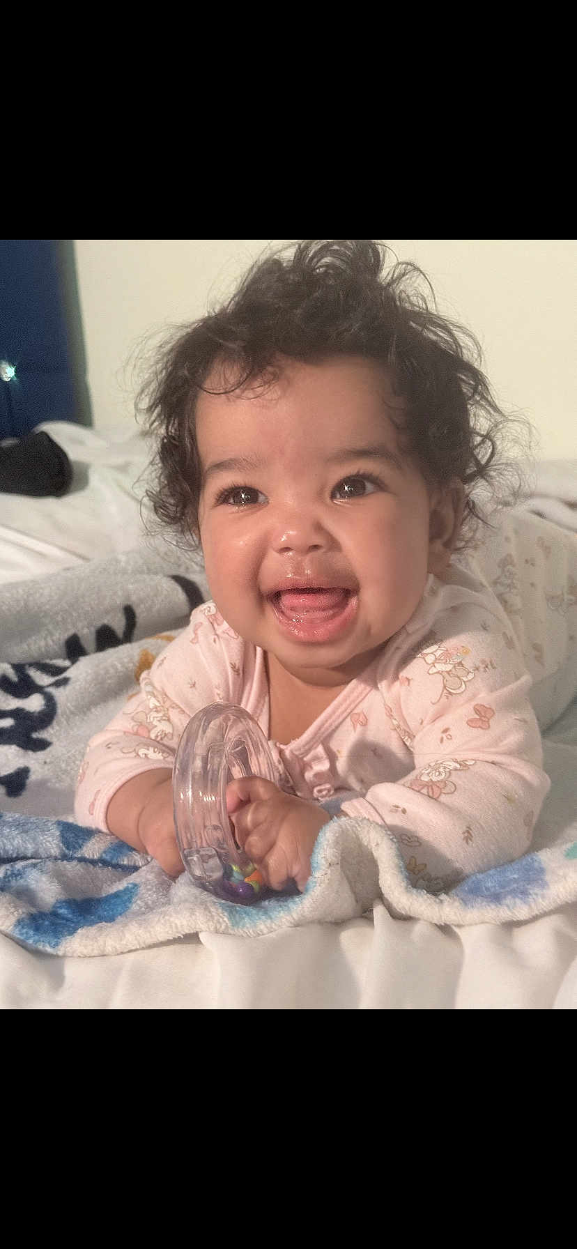 Alani joined the competition — help win amazing prizes! baby, infant, smiling, curly_hair, toy, blanket, bed, pajamas, tongue_out, happy, bright_eyes, hands, rattle, cozy, portrait, skin, indoor, cute, lying_down, nursery