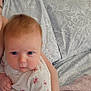 baby, infant, child, person, face, blue_eyes, clothing, floral_pattern, onesie, bed, pillow, blanket, adult_arm, indoor, portrait, close_up, skin, hair, expression, holding