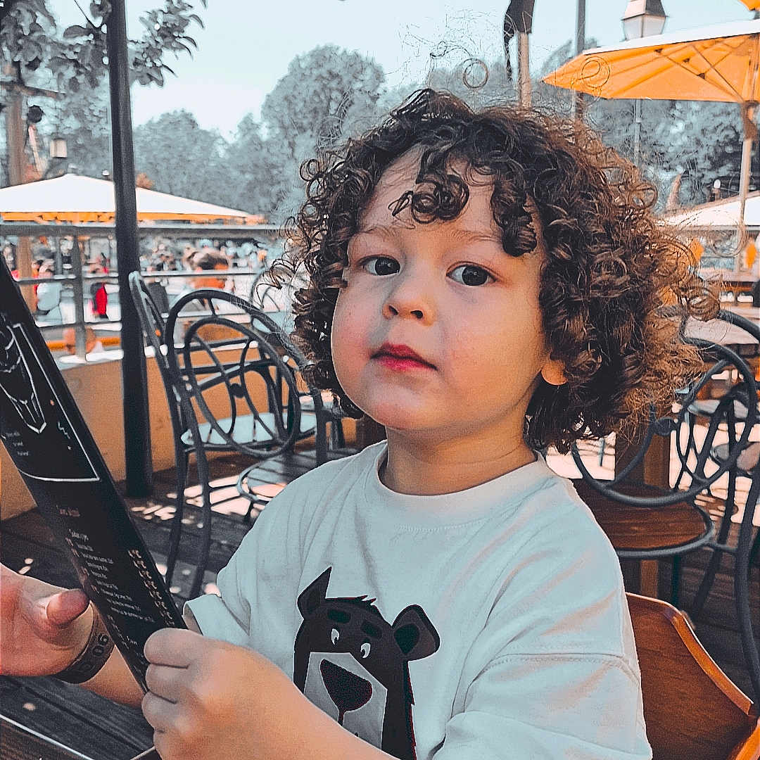 Eden a rejoint le concours — aidez-le/la à gagner de superbes lots ! bear_graphic, chair, child, curious, curly_hair, cute, daylight, menu, metal_chair, outdoor, park, person, restaurant, seat, summer, toddler, umbrella, white_tshirt, wooden_table, yellow_umbrella