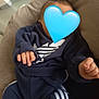 baby, blue_heart_emoji, cushion, infant, navy_blue_clothing, soft_texture, sitting, hand, fingers, indoor, casual_wear, tracksuit, child, comfort, relaxation, small_hands, fabric, person, young_child, hidden_face