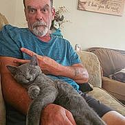 Fuzzy is registered to the contest to win money with this photo: man, cat, gray_cat, couch, living_room, blue_shirt, sitting, relaxed, pet, tattoo, facial_hair, wall_art, flower_vase, can, furniture, home_interior, animal, portrait, indoor, resting